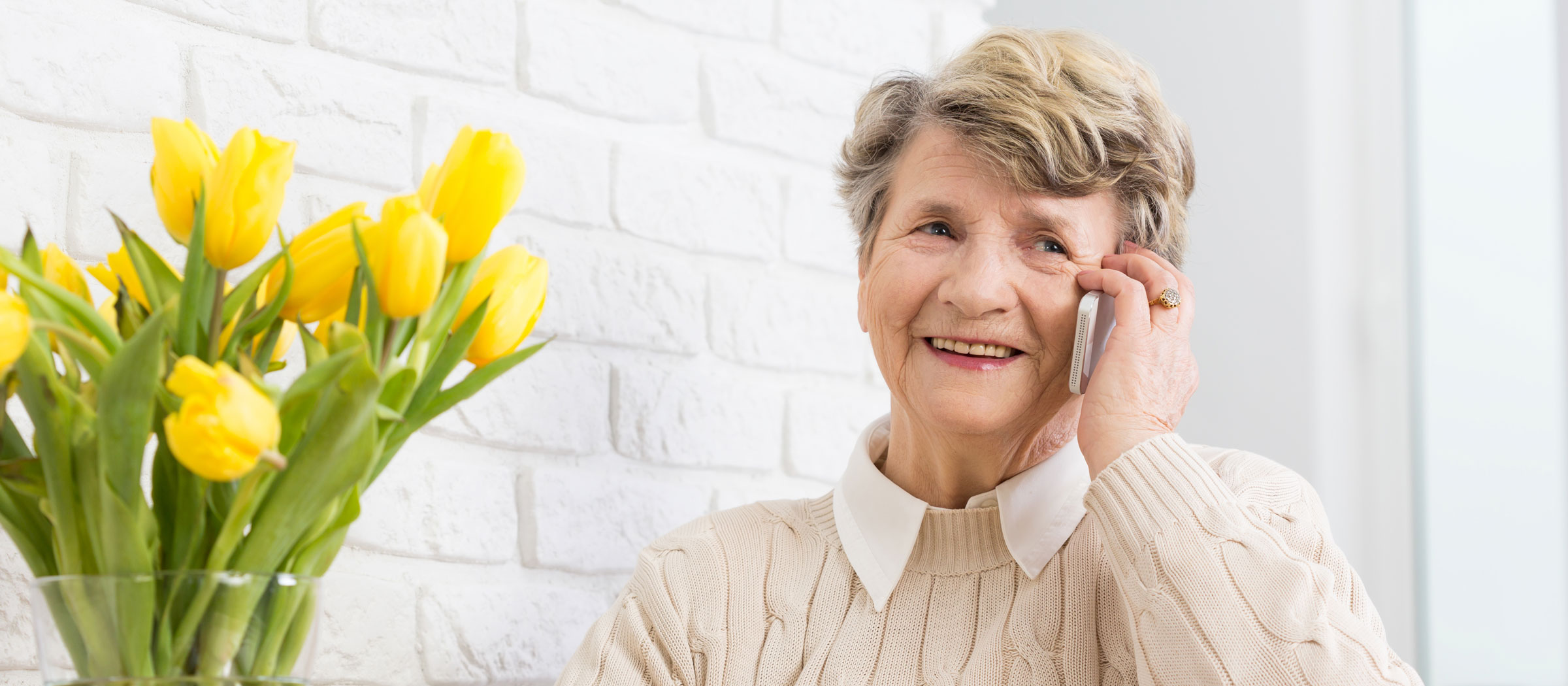 An older woman smiling on the phone beside a vase of yellow tulips.