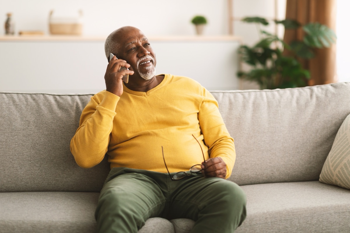 An older man in a yellow sweater smiling warmly while on the phone.